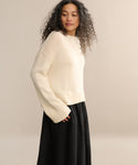 A woman with long, curly hair wears the cream-colored Cameron Crewneck with wide sleeves and a black skirt, standing against a plain, light beige background.