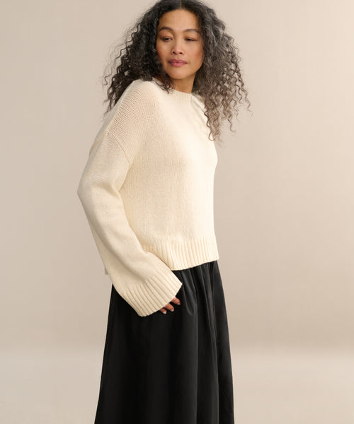 A woman with long, curly hair wears the cream-colored Cameron Crewneck with wide sleeves and a black skirt, standing against a plain, light beige background.