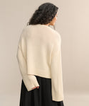 A woman with long, curly dark hair is seen from behind wearing the Cameron Crewneck, an oversized cream-colored sweater, paired with a black skirt against a plain beige background.