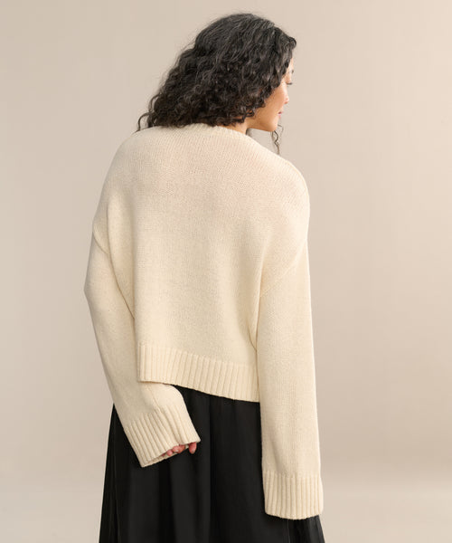 A woman with long, curly dark hair is seen from behind wearing the Cameron Crewneck, an oversized cream-colored sweater, paired with a black skirt against a plain beige background.