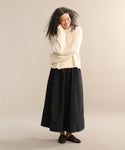 A person with long curly hair smiles with arms crossed, wearing the cream Cameron Crewneck, a long black skirt, and black slip-on shoes against a plain beige background.