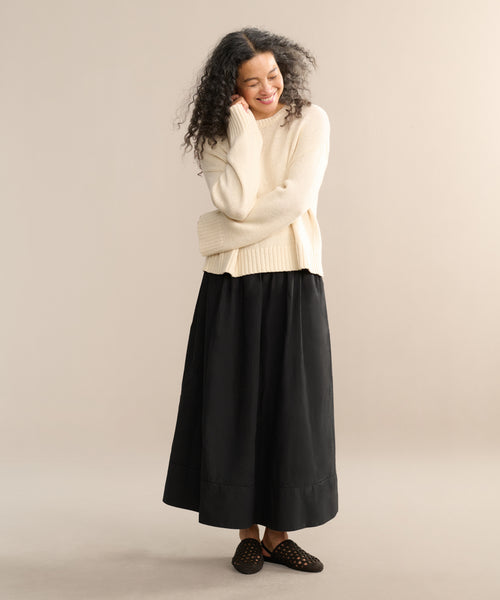 A person with long curly hair smiles with arms crossed, wearing the cream Cameron Crewneck, a long black skirt, and black slip-on shoes against a plain beige background.
