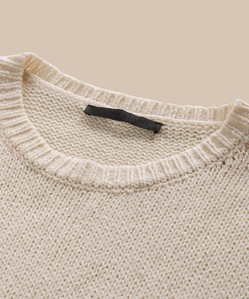 Close-up of the Cameron Crewneck, a cream-colored sweater with a ribbed neckline and black label inside the collar, shown against a beige background—a lightweight piece ideal for layering.
