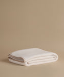 A folded cream-colored knit blanket with a beige fringed edge, displayed on a solid beige background.