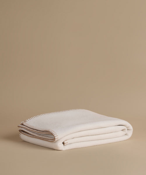 A folded cream-colored knit blanket with a beige fringed edge, displayed on a solid beige background.
