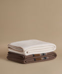 Two folded woolen throws stacked on a beige background. The top throw is off-white with a cream border, and the bottom throw is brown with a subtle plaid pattern.