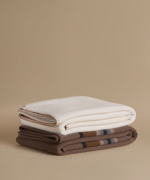 Two folded woolen throws stacked on a beige background. The top throw is off-white with a cream border, and the bottom throw is brown with a subtle plaid pattern.