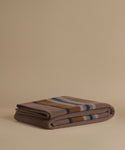 A folded wool blanket with a classic stripe pattern in taupe, camel, navy, and cream, displayed on a solid beige background.