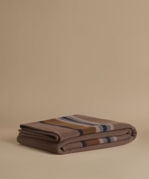 A folded wool blanket with a classic stripe pattern in taupe, camel, navy, and cream, displayed on a solid beige background.