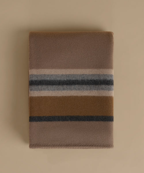 A folded wool blanket with a classic stripe pattern in taupe, camel, navy, and cream, displayed on a solid beige background.