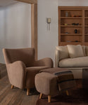 A cozy interior scene showing a textured taupe armchair paired with a matching ottoman and a folded throw blanket, set in a living room with a sofa, wooden shelving, and wood flooring.