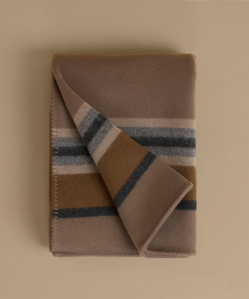 A folded wool blanket with a classic stripe pattern in taupe, camel, navy, and cream, displayed on a solid beige background.