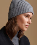 A close-up side profile of a woman wearing a ribbed knit gray beanie and a dark coat against a warm beige background.