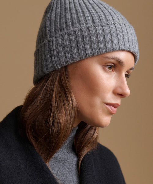 A close-up side profile of a woman wearing a ribbed knit gray beanie and a dark coat against a warm beige background.