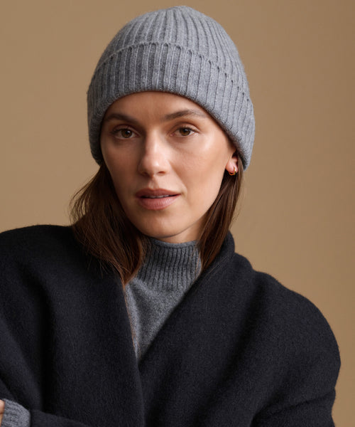 A woman wearing a light gray ribbed knit beanie and a dark charcoal wrap cardigan over a gray turtleneck, set against a solid warm beige background.