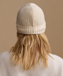 A person viewed from behind wearing a cream ribbed knit beanie and a matching cream sweater against a solid beige background.