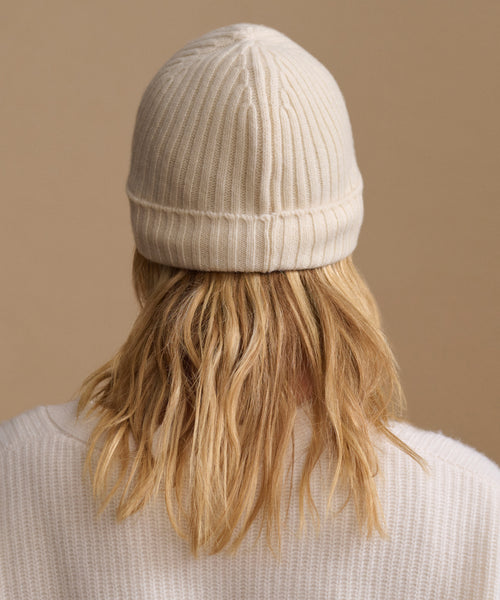 A person viewed from behind wearing a cream ribbed knit beanie and a matching cream sweater against a solid beige background.