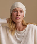 A woman wearing a cream ribbed knit beanie and matching cream knit top against a warm beige studio background.