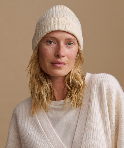 A woman wearing a cream ribbed knit beanie and a matching cream knit top, photographed against a warm beige studio background.