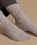 Close-up of a person wearing ribbed knit ankle socks in a heathered taupe-beige color, with dark leggings visible at the top, against a solid tan background.