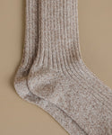 A close-up of a pair of ribbed knit socks in a heathered taupe-brown and cream colorway, displayed against a solid beige background.