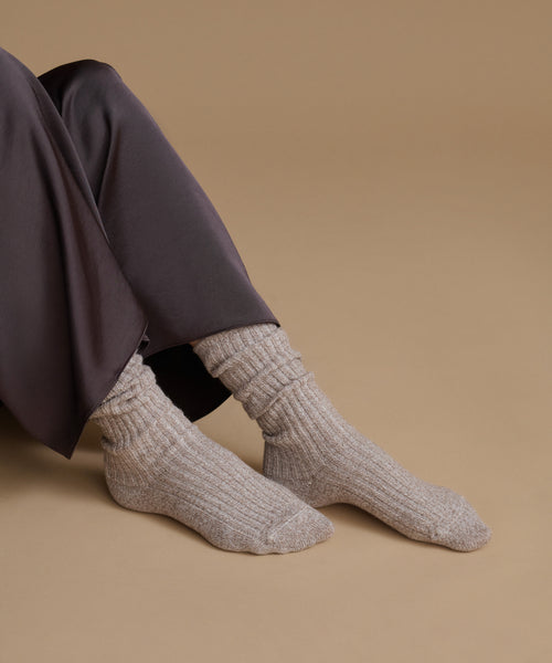 A person wearing ribbed ankle socks in a light taupe-beige color, paired with dark charcoal trousers, posed against a solid warm tan background.