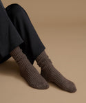 A close-up of a person's feet wearing dark brown ribbed knit socks with black trousers, set against a solid warm beige background.