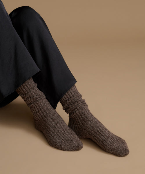 A close-up of a person's feet wearing dark brown ribbed knit socks with black trousers, set against a solid warm beige background.