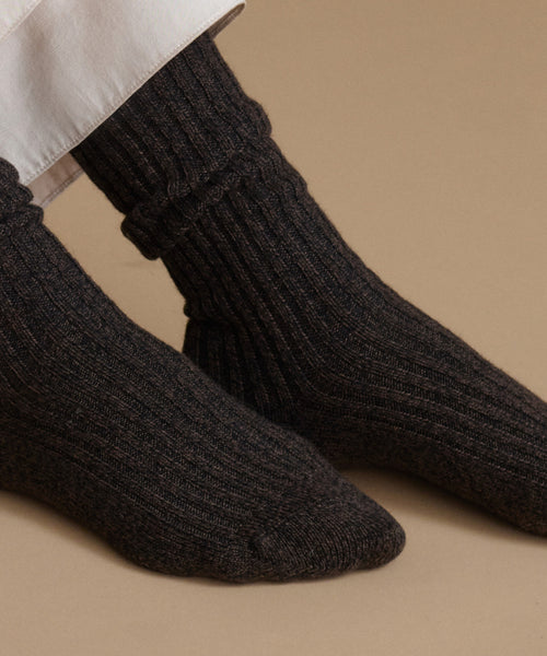 A close-up of a person's feet wearing dark charcoal gray ribbed knit socks, paired with a white shirt cuff, set against a solid beige background.