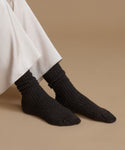 A close-up of a person's feet wearing dark charcoal gray ribbed knit socks, with the hem of a white dress or skirt visible at the top, set against a solid beige background.