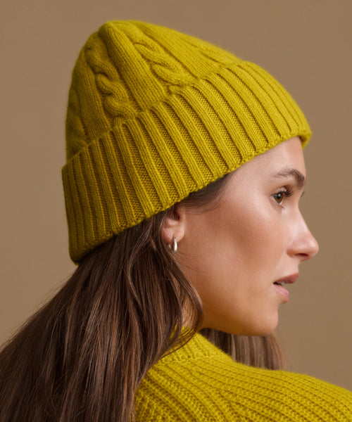 A woman in profile wearing a mustard yellow cable-knit beanie and a matching knit sweater against a solid beige background.