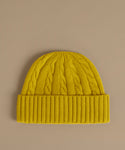 A bright yellow cable-knit beanie hat with a ribbed cuff, displayed flat against a solid taupe background.