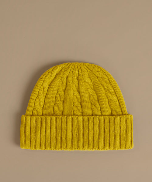 A bright yellow cable-knit beanie hat with a ribbed cuff, displayed flat against a solid taupe background.