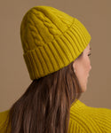 A woman shown in side profile wearing a mustard yellow cable-knit beanie and a matching knit sweater against a plain warm beige background.