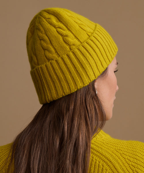 A woman shown in side profile wearing a mustard yellow cable-knit beanie and a matching knit sweater against a plain warm beige background.
