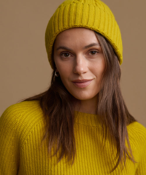 A woman wearing a bright yellow ribbed knit beanie and matching yellow knit sweater, photographed against a warm beige studio background.
