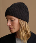 A woman wearing a dark charcoal gray cable-knit beanie hat, shown in profile against a solid warm brown background.