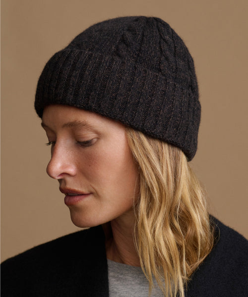 A woman wearing a dark charcoal gray cable-knit beanie hat, shown in profile against a solid warm brown background.