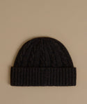 A dark charcoal cable-knit beanie with a ribbed cuff, displayed flat against a solid beige background.