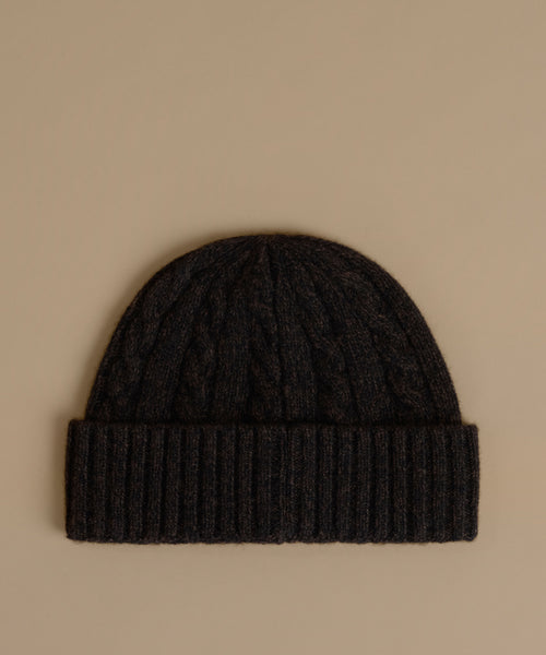 A dark charcoal cable-knit beanie with a ribbed cuff, displayed flat against a solid beige background.