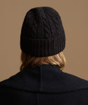 Back view of a person wearing a black cable-knit beanie and a dark coat against a solid tan background.