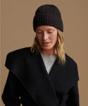 A woman wearing a black cable-knit beanie and a black wrap coat over a light gray top, photographed against a warm tan studio background.