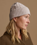 A woman in profile wearing a light taupe cable-knit beanie and a brown coat, set against a solid warm beige background.