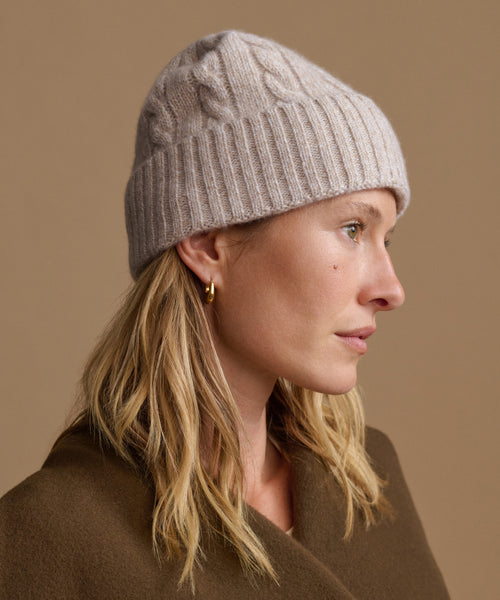 A woman in profile wearing a light taupe cable-knit beanie and a brown coat, set against a solid warm beige background.