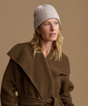 A woman wearing a chunky cable-knit beige beanie and a brown wrap coat with a wide collar and tie belt, posed against a solid warm brown background.