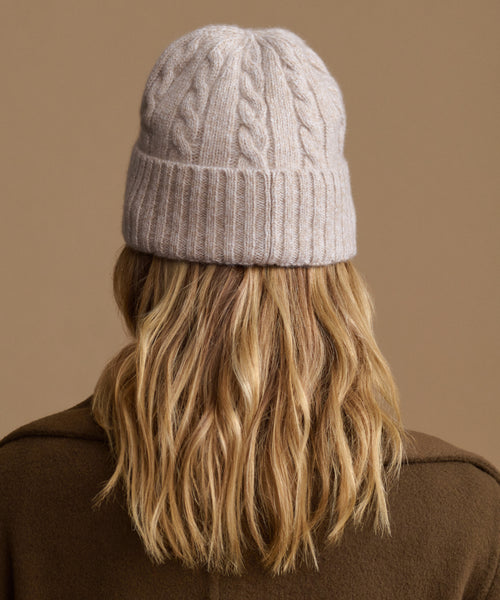 A person shown from the back wearing a light beige cable-knit beanie and a brown coat, against a solid warm tan background.