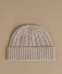 A beige cable-knit beanie with a ribbed cuff, photographed flat against a solid beige background.