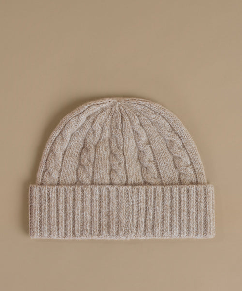 A beige cable-knit beanie with a ribbed cuff, photographed flat against a solid beige background.