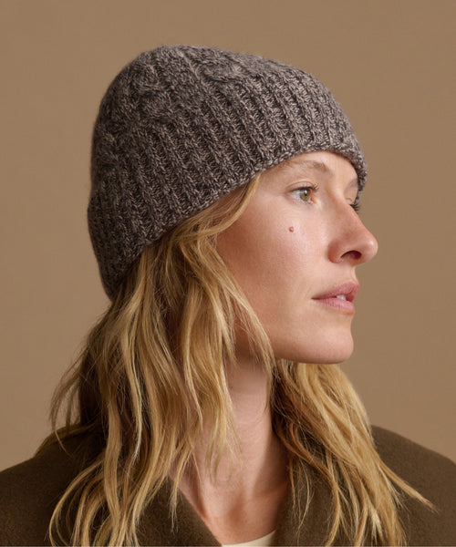 A woman in profile wearing a dark gray marled knit beanie hat, set against a solid warm beige background.