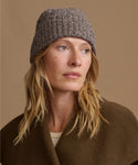 A woman wearing a textured gray knit beanie and a brown wool coat, photographed against a solid warm beige background.